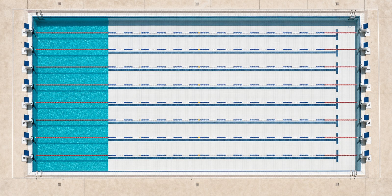 Aerial view of an Olympic swimming pool. The leftmost portion is filled with turquoise water and represents 129,000 gallons; the remaining 80 percent of the pool is completely dry, showing bare white tile.